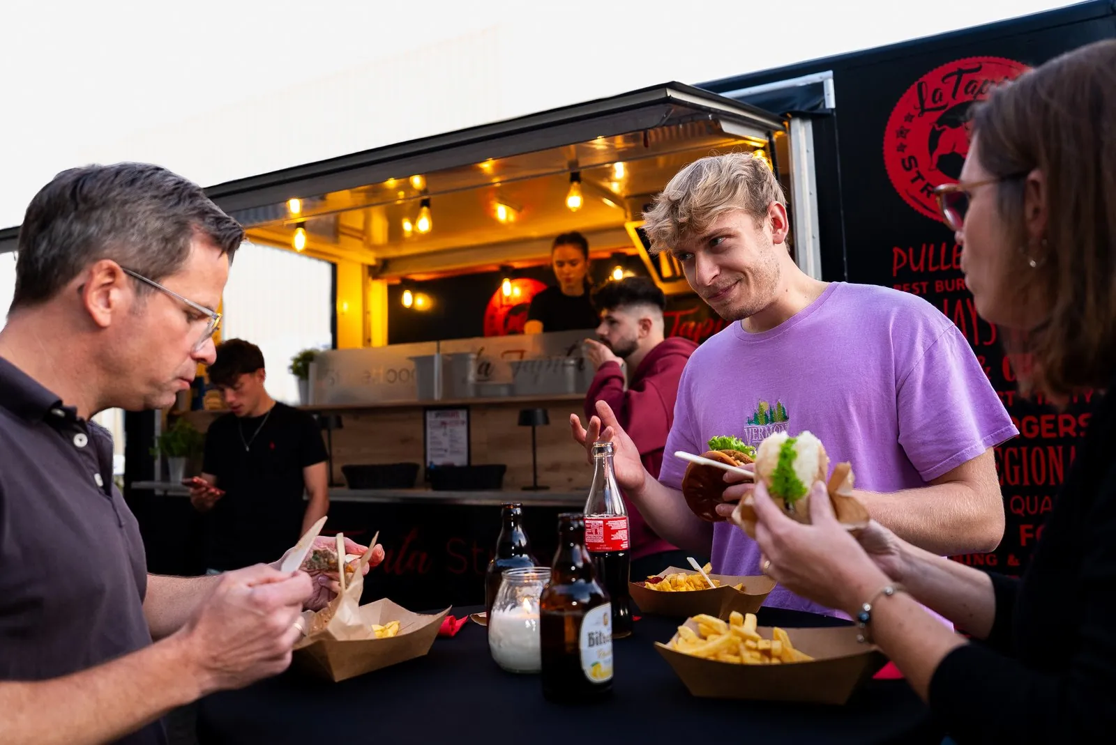 Gäste mit Burgern und Pommes am Stehtisch vor dem Foodtrailer am Abend