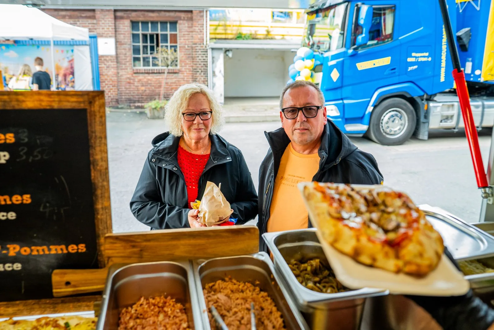 Pinsa wird an der Foodtruck-Theke an zwei Gäste ausgegeben, im Hintergrund ein Firmengelände mit Luftballons